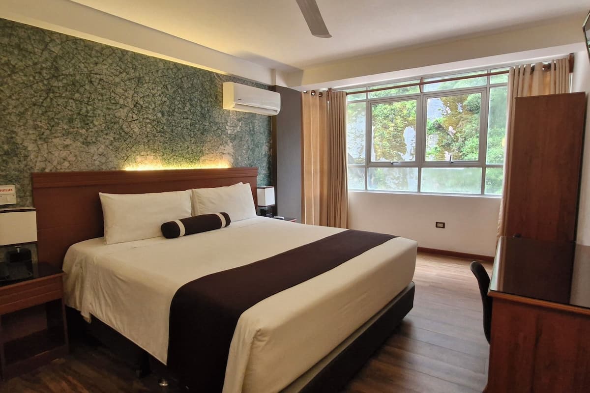 A hotel room with a bed, nightstand, lamp, air conditioning unit, window with curtains, and a TV mounted on the wall.
