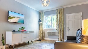 A living room with a flat-screen TV mounted on the wall, a white console table, a floor lamp, a sofa, and a window with curtains. - Beachside All Suites Hotel (Miami Beach)