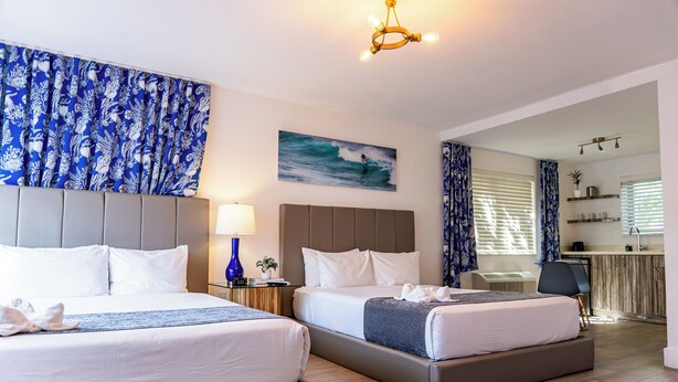 A hotel room with two beds, a large painting of a surfer, a chandelier, and a view into the kitchen area. - Beachside All Suites Hotel (Miami Beach)