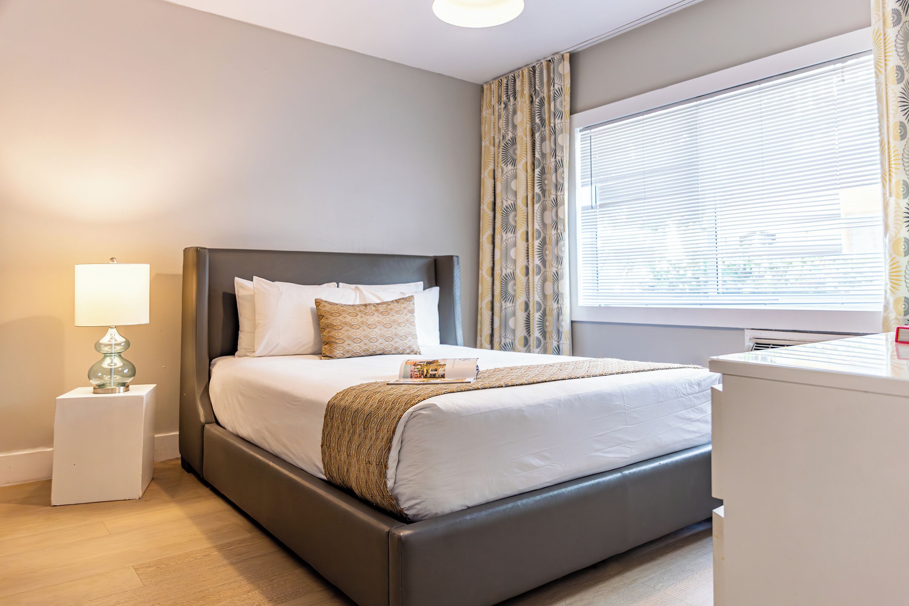 Un dormitorio moderno con cama, mesita de noche, lámpara y ventana con cortinas.
