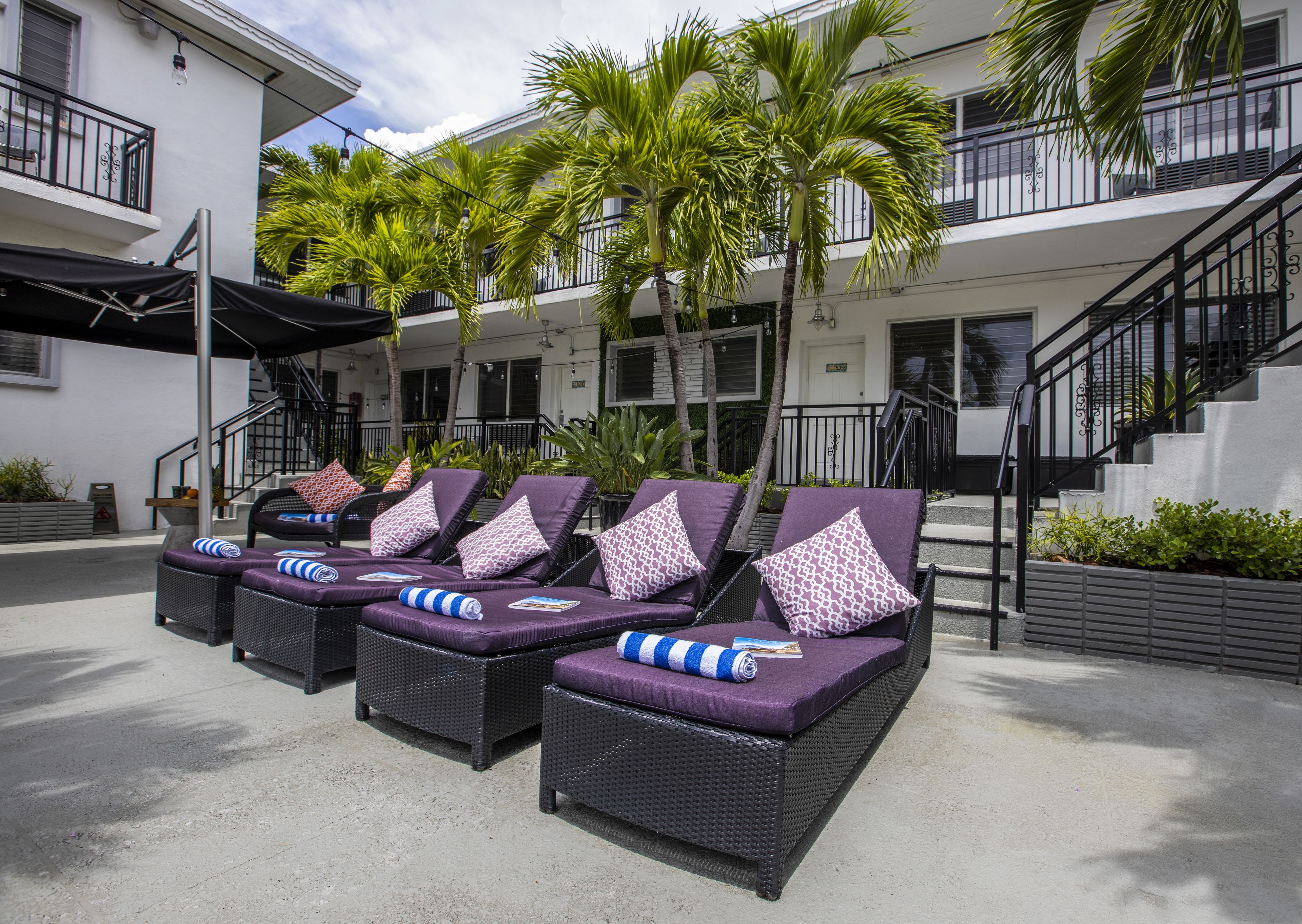 Outdoor lounge chairs with purple cushions and patterned pillows, arranged in front of a building with a white exterior and black railings.