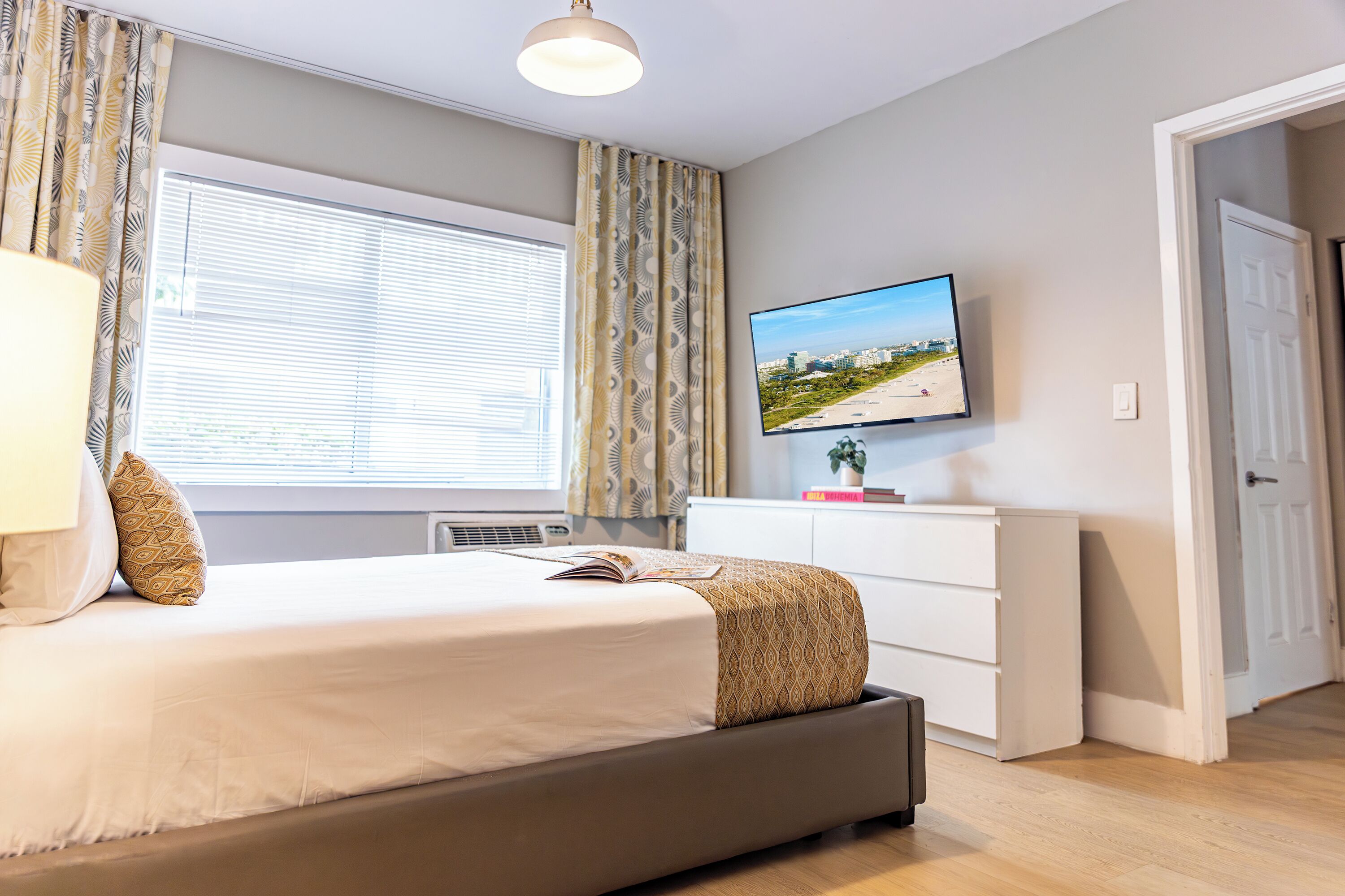 A bedroom with a bed, a TV on the wall, a window with curtains, and a door.