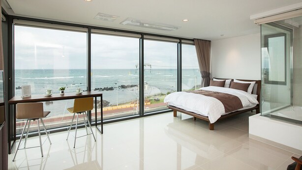 A modern bedroom with a large window offering a sea view, a bed, a desk with a chair, and a glass partition. - Vista View Resort (Jeju City)