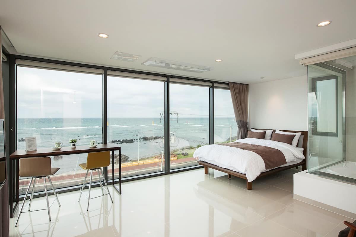 Un dormitorio moderno con un ventanal que ofrece vistas al mar, una cama, un escritorio con silla y un separador de vidrio.