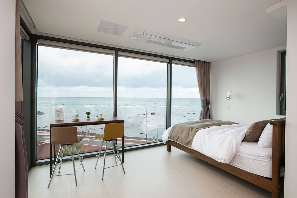 Habitación de hotel moderna con un ventanal que ofrece vistas al mar. Tiene una cama con sábanas blancas, un escritorio con silla, y una mesita con una planta y una taza.