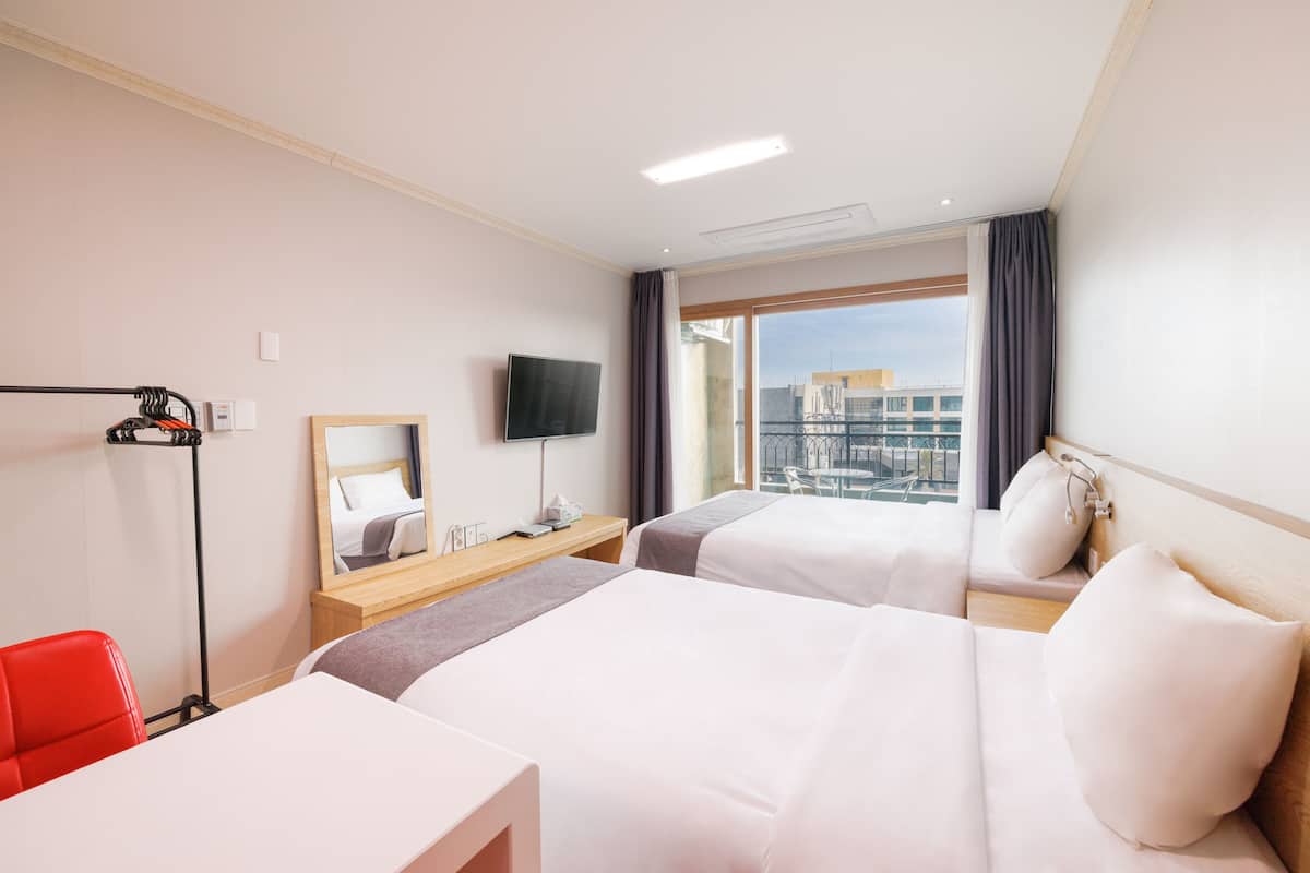 A hotel room with two beds, a desk, a chair, a TV, and a large window with a city view.
