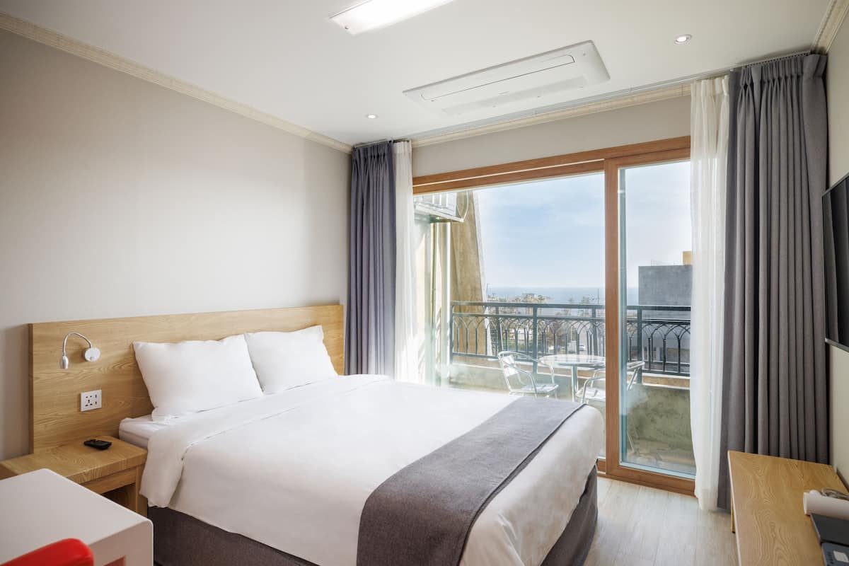 A hotel room with a large bed, a desk, a chair, a TV, and a balcony with a view.