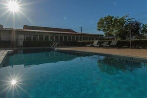 Una piscina azul cristalina con el sol brillando directamente arriba, rodeada por una zona vallada con sillas de descanso.