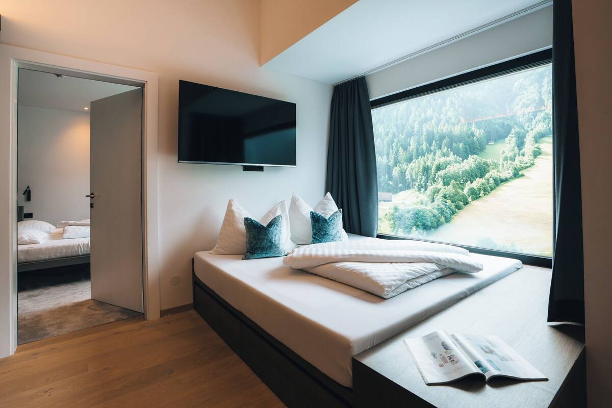 A modern bedroom with a large window, a flat-screen TV, and a bed with pillows.