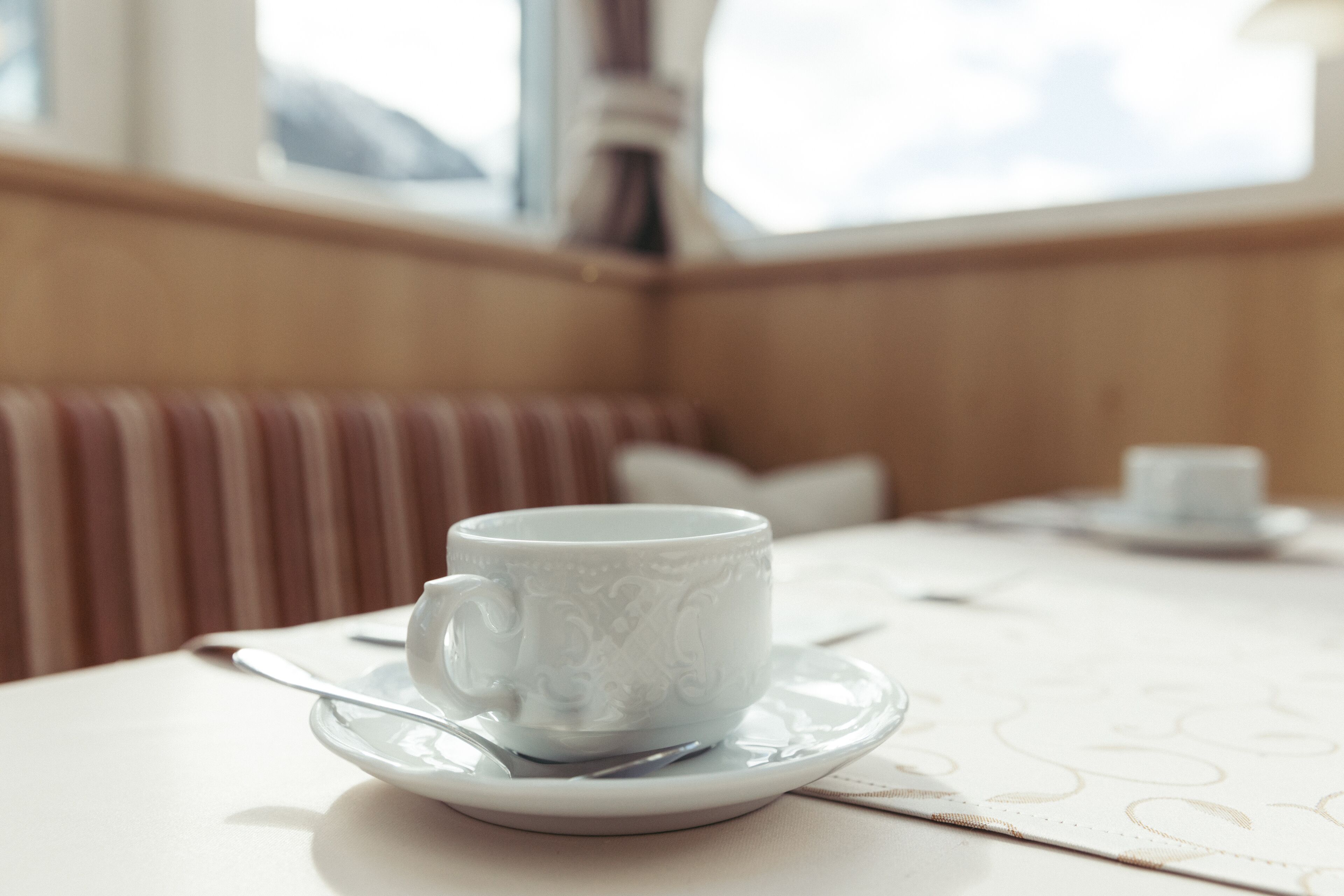 A white cup with a saucer and spoon on a table in a room with wooden paneling and a window.
