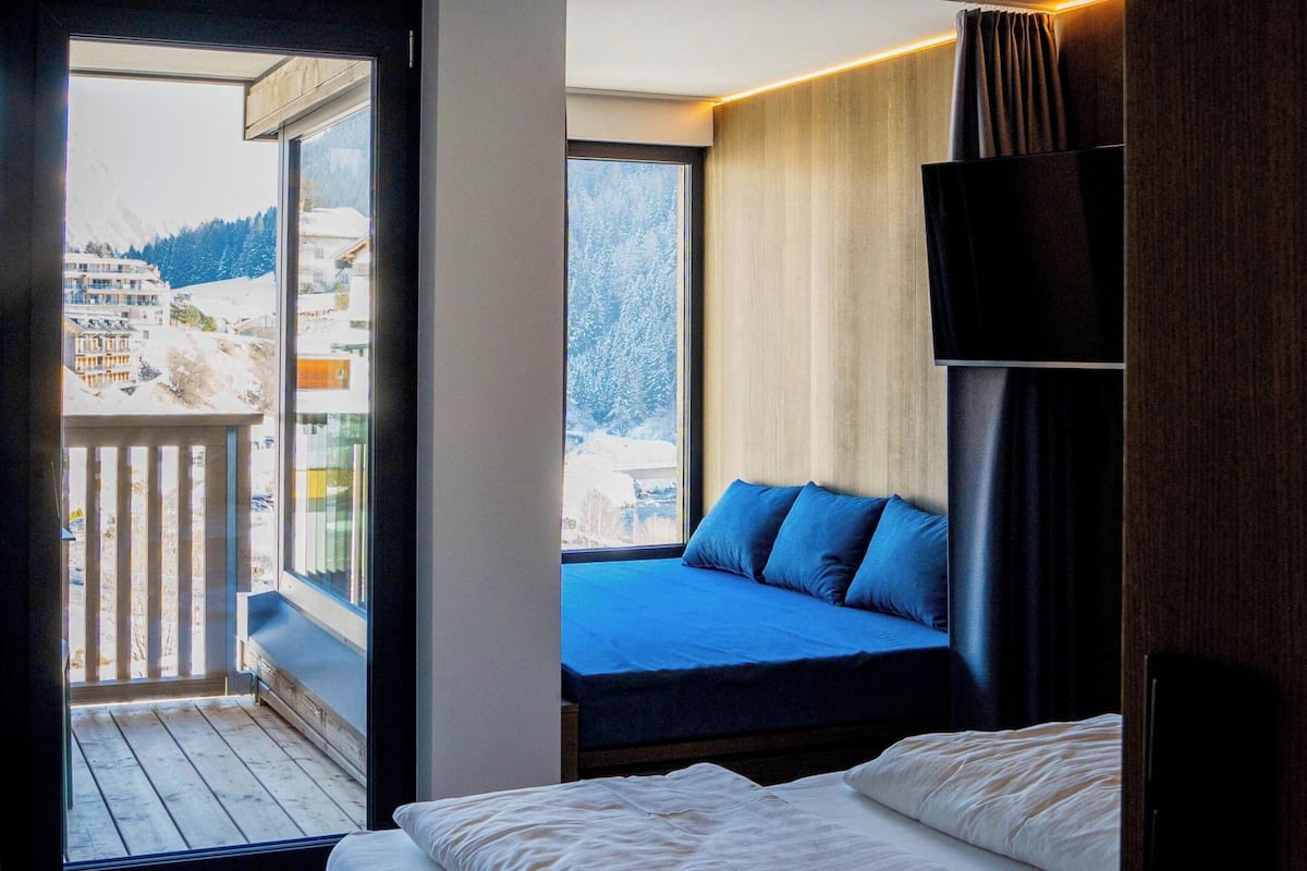 A bedroom with a bed, a bench, a TV, and a balcony with a view of buildings and trees.