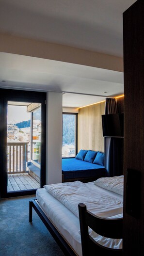 A bedroom with a bed, a bench, a TV, and a balcony with a view of buildings and trees.