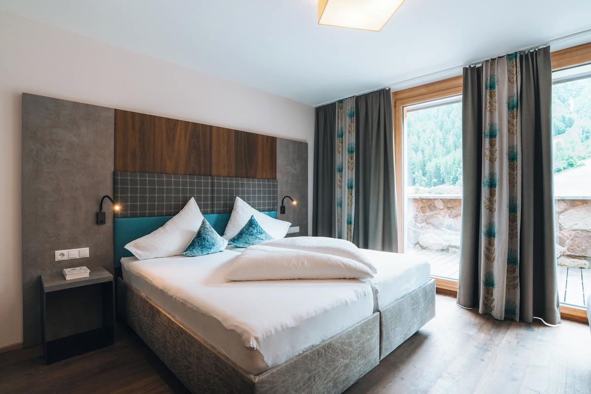 A modern hotel room with a large bed, a nightstand, a wall-mounted lamp, and a view of a forested area.
