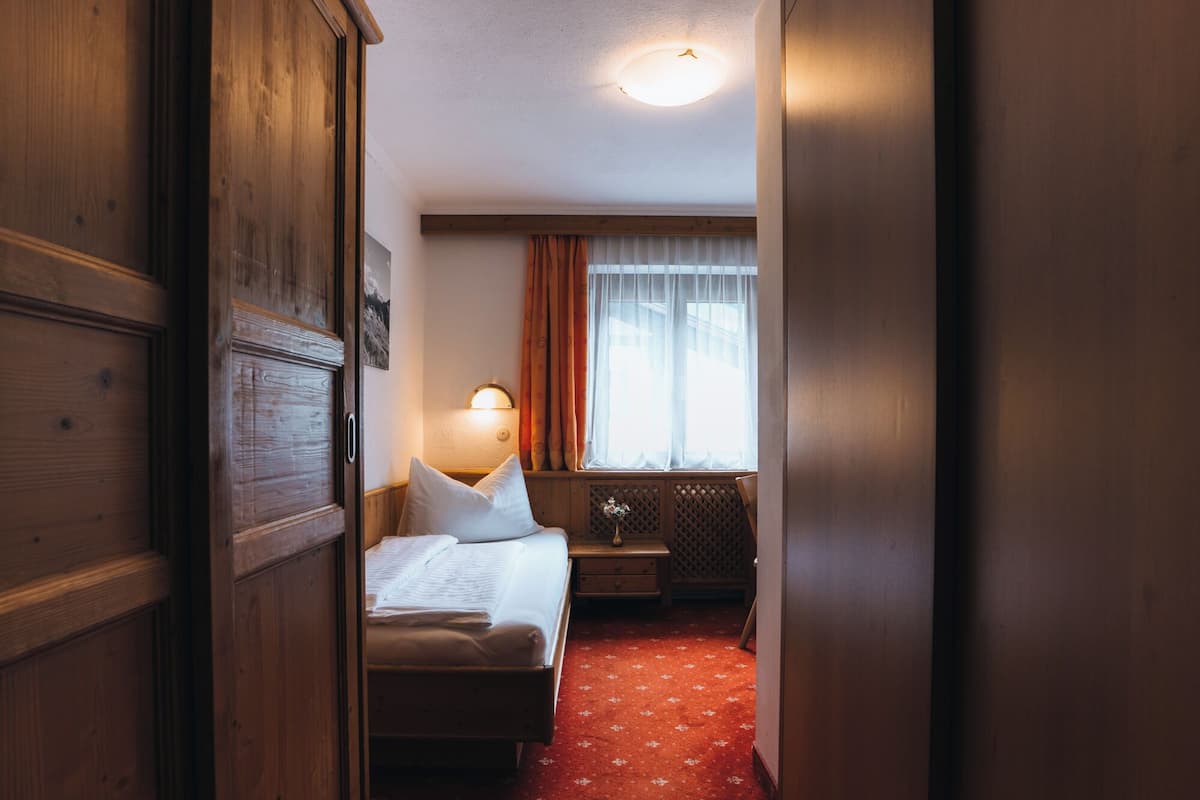 A hotel room with a wooden wardrobe, a bed with white linens, a nightstand with a lamp, and a window with curtains.