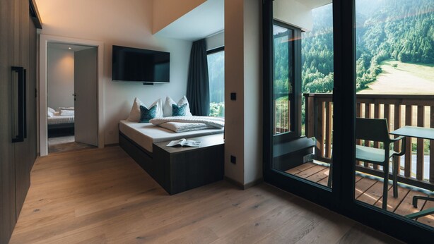 A modern bedroom with a large bed, a TV, and a balcony with a view of a forest.
