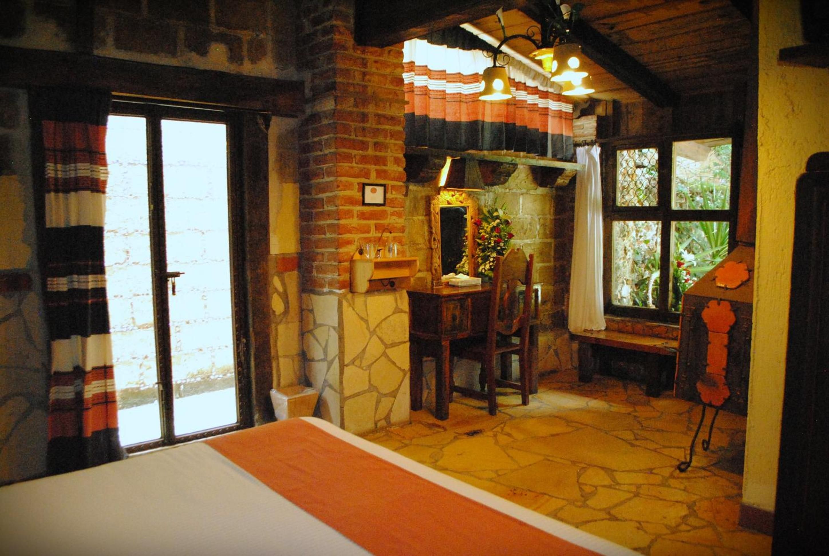 A room with a bed, a table with chairs, a window with curtains, and a stone wall.