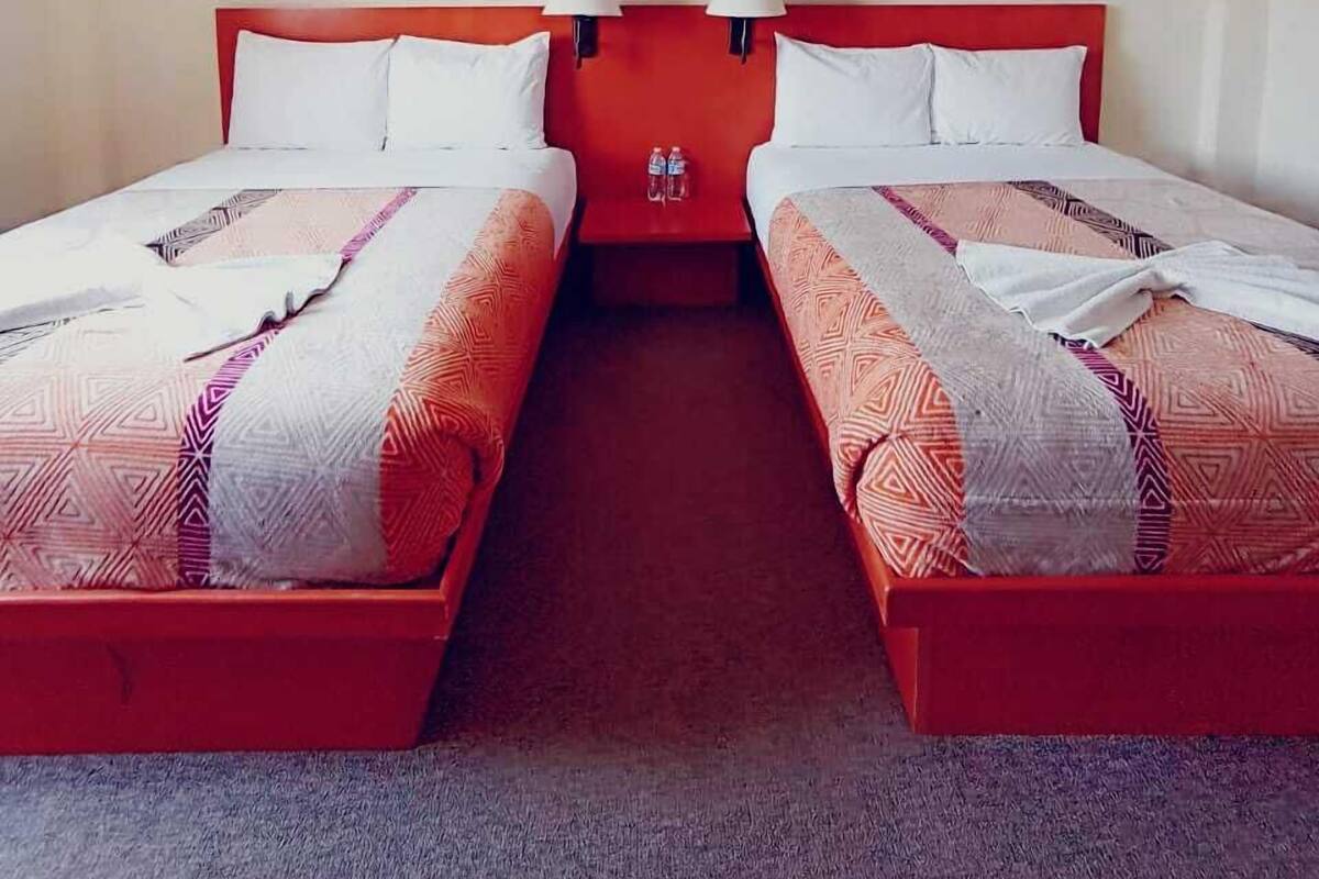 A hotel room with two beds, a red headboard, a painting of a city skyline, and a nightstand with two lamps.
