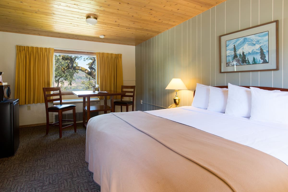 A hotel room with a large bed, a desk with chairs, a lamp, and a painting on the wall.