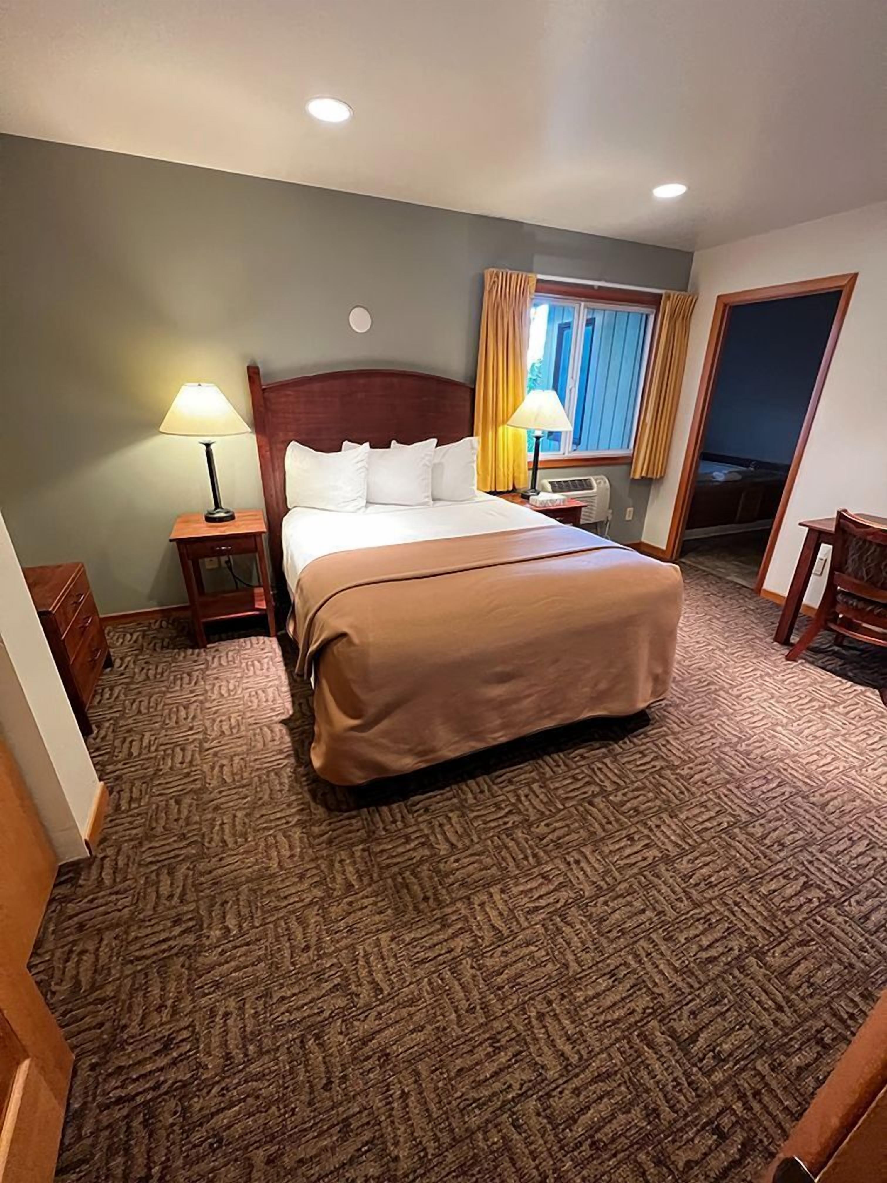 Une chambre d’hôtel avec un grand lit, une table de chevet avec une lampe, une fenêtre avec des rideaux et une porte donnant sur une autre pièce.
