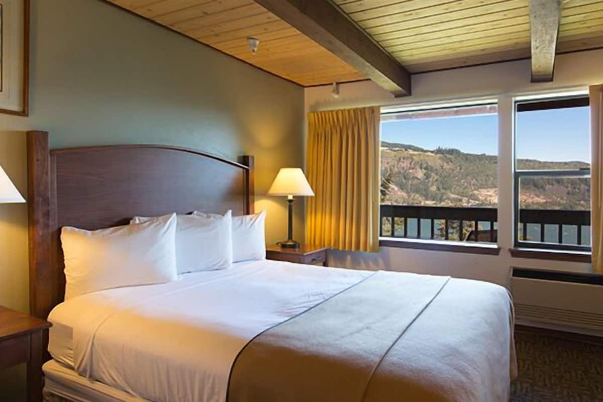 A hotel room with a bed, two bedside lamps, a window with a view of the outdoors, and a wooden ceiling.