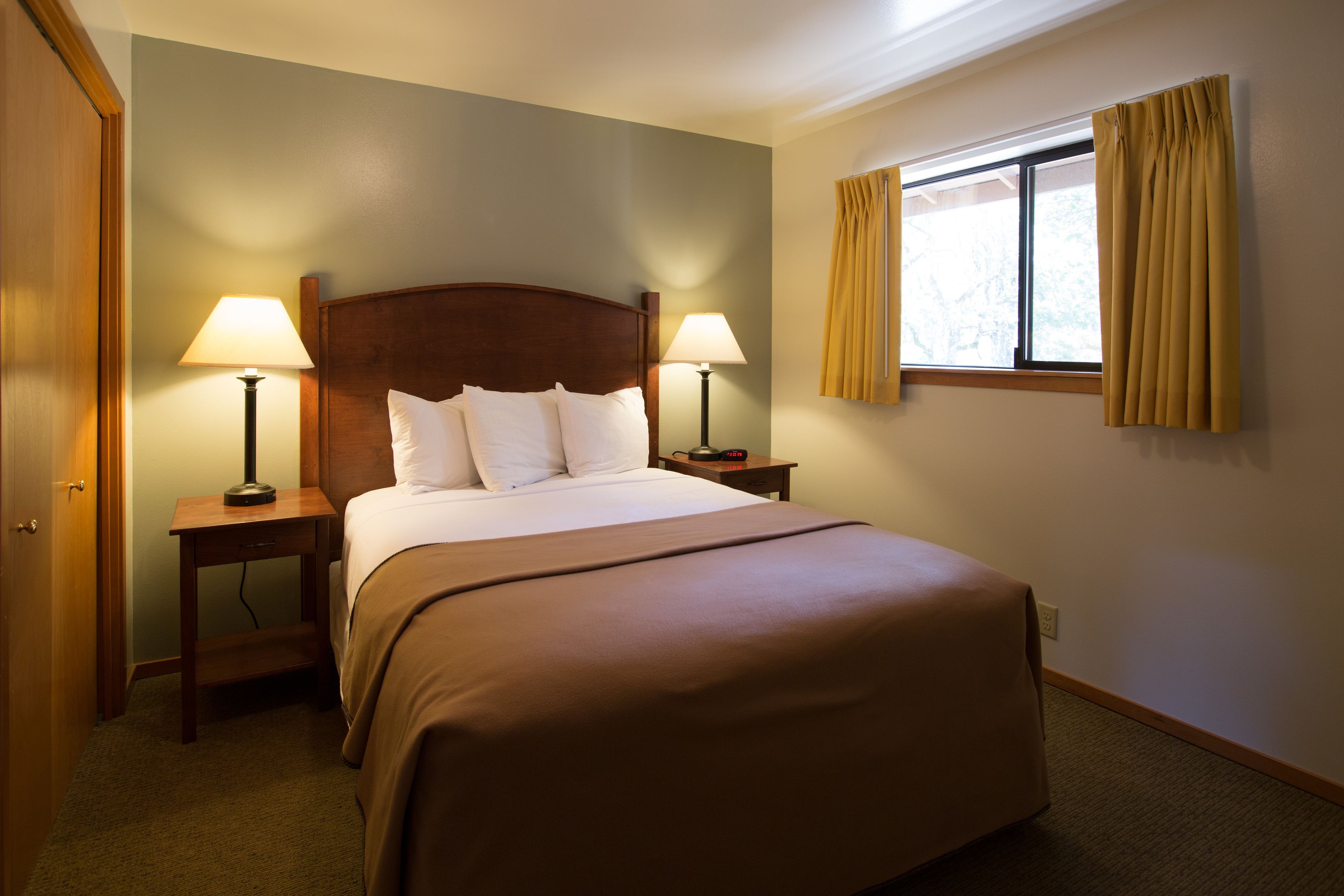 Habitación de hotel con una cama grande, dos mesitas de noche con lámparas, una ventana con cortinas y una puerta.