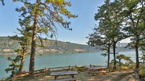 Un área de picnic junto al lago con bancos y una cerca de madera, rodeada de árboles y colinas.