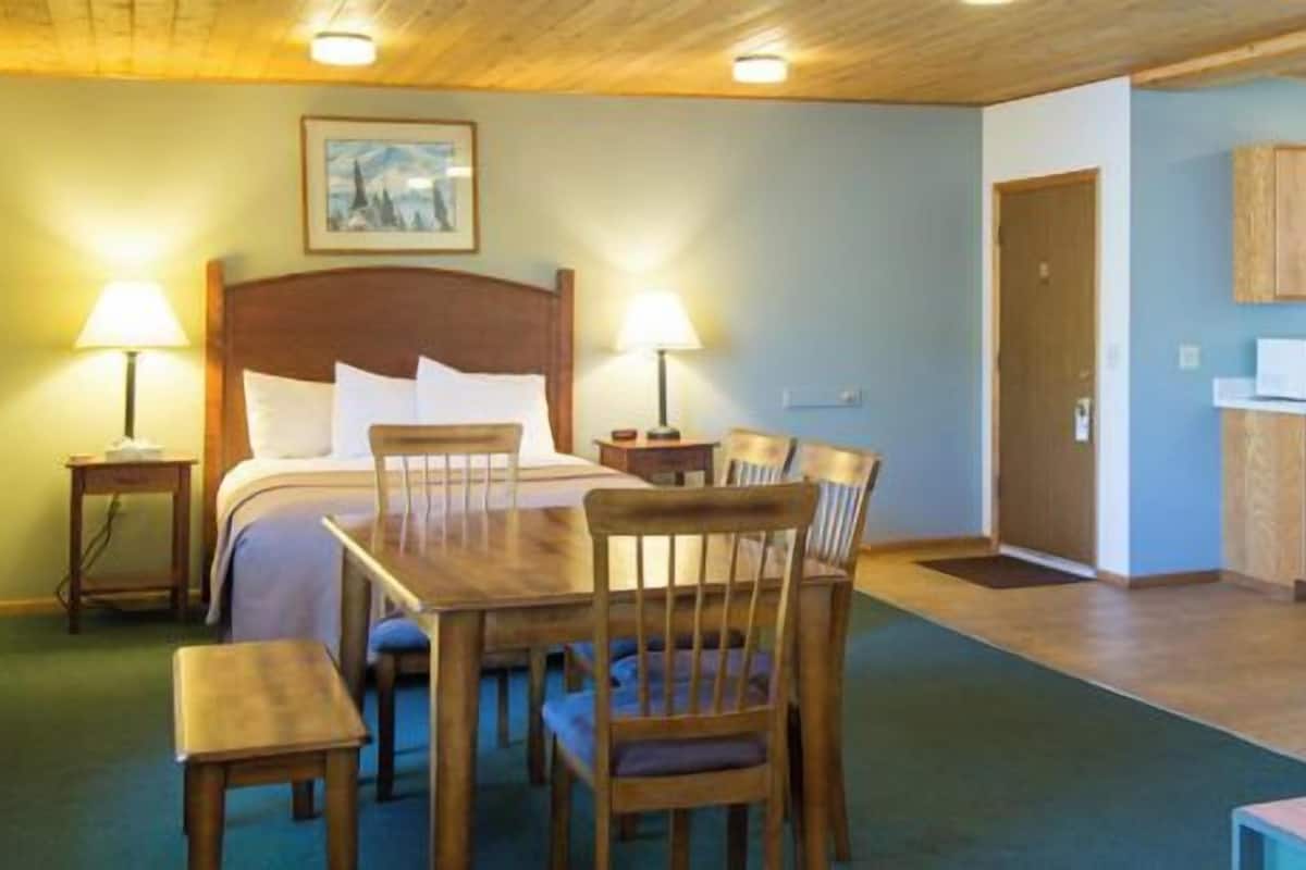 A hotel room with a bed, two bedside tables with lamps, a wooden dining table with chairs, a kitchenette with a microwave, and a blue sofa.