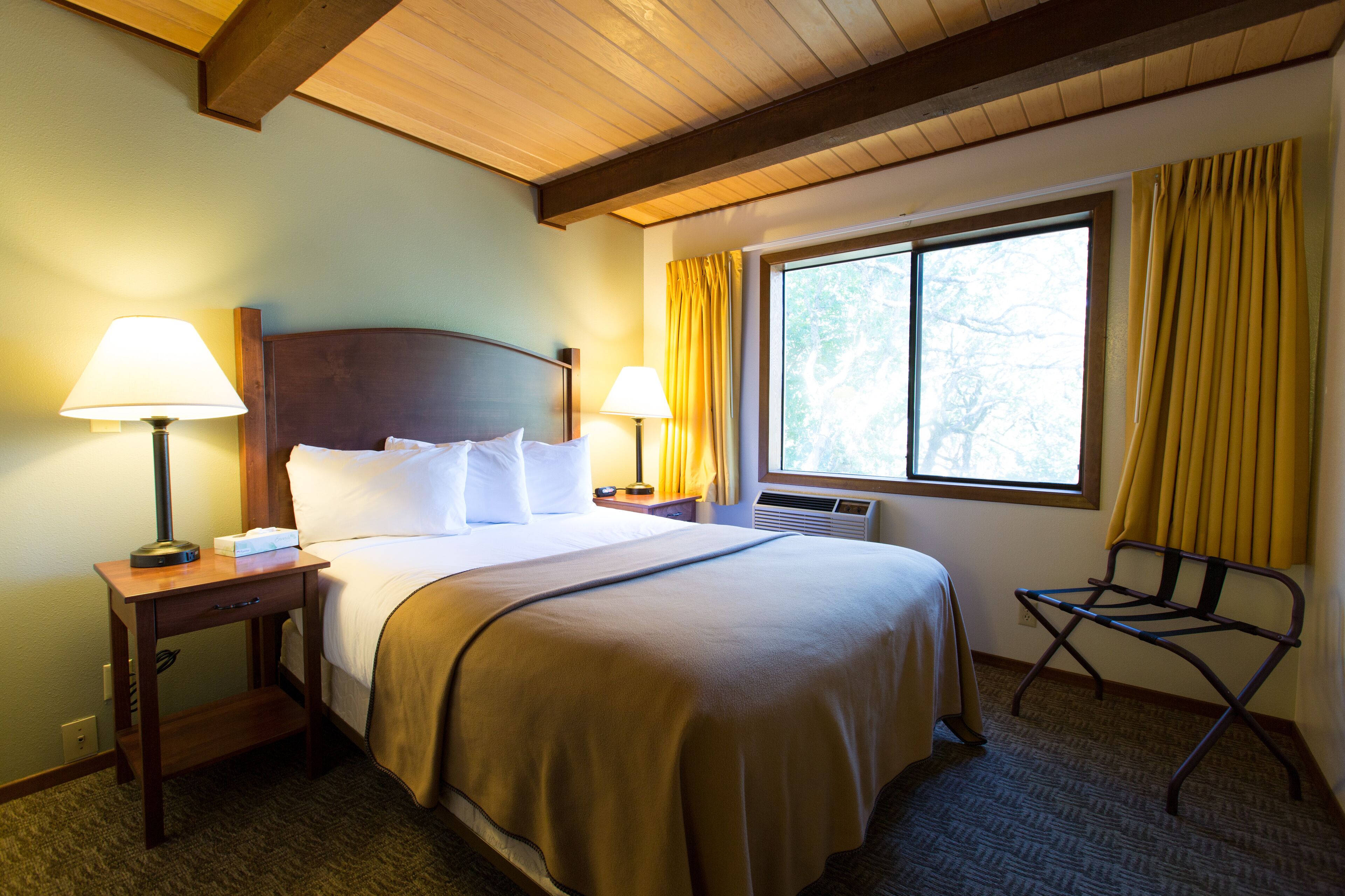 Een hotelkamer met een groot bed, twee bedlampen, een raam met gordijnen, een stoel en een houten plafond.