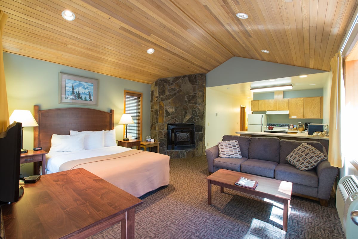 A hotel room with a bed, a sofa, a coffee table, a TV, a fireplace, and a kitchen area.