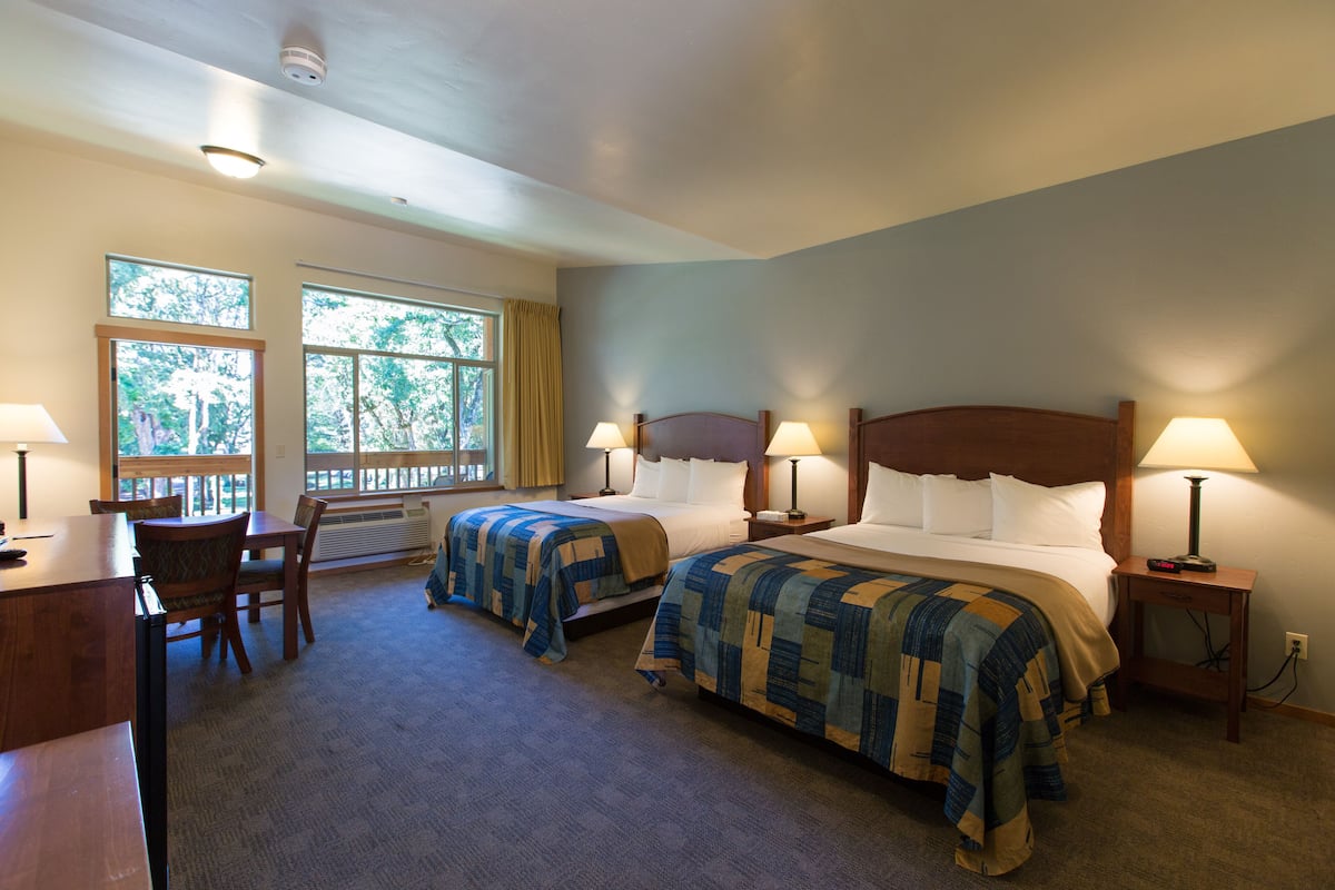 A hotel room with two beds, a desk, and a chair. There are windows with curtains, a heater, and a lamp on the nightstand.