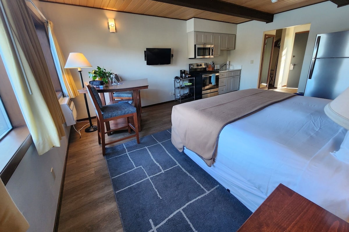 A hotel room with a bed, a desk with a chair, a TV, a kitchenette, and a window with curtains.