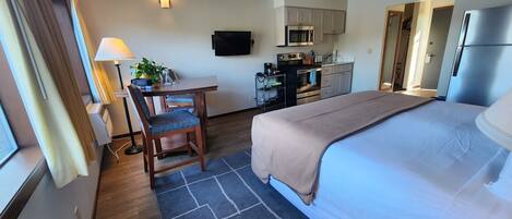 Ein Hotelzimmer mit Bett, Schreibtisch mit Stuhl, Fernseher, Küchenzeile und einem Fenster mit Vorhängen.