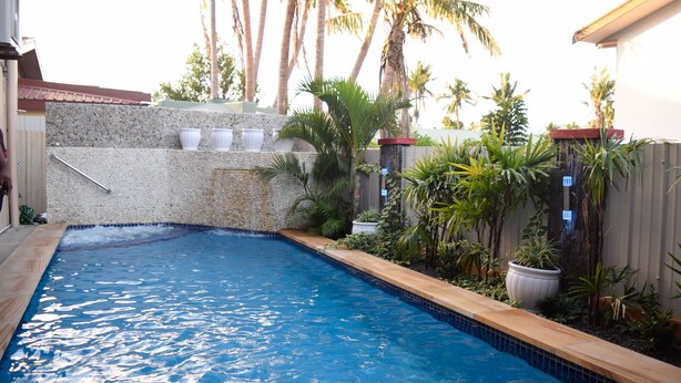 A swimming pool with a pebble-lined edge, surrounded by tropical plants and a stone wall. - Al-Minhaj Service Apartments (Nadi)