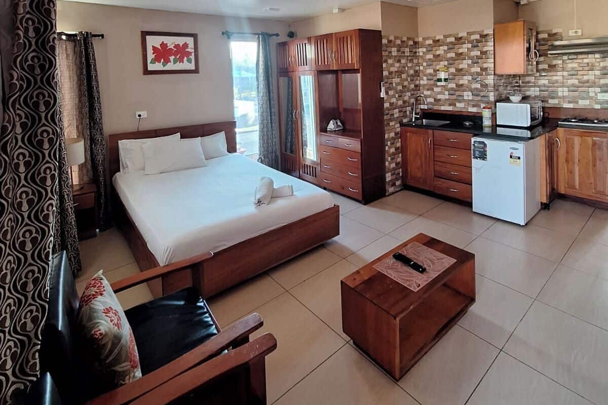 A hotel room with a bed, a wooden chair, a small table, a kitchenette, and a bathroom with a shower.