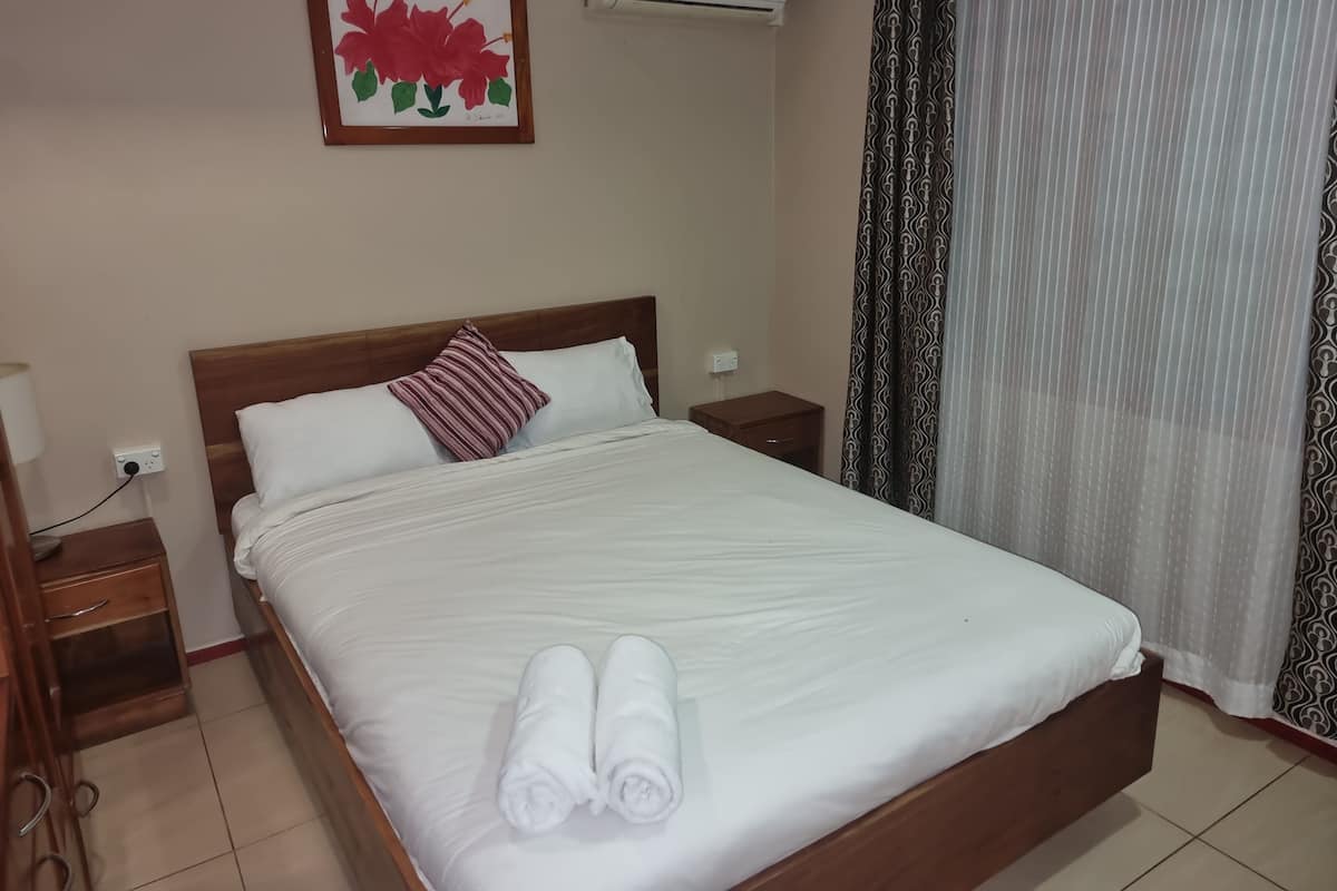 A neatly made bed with white linens, a wooden headboard, a bedside table with a lamp, and a wall-mounted air conditioner.