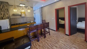 A modern kitchen with a central island, wooden dining table, and chairs. There is a separate bedroom with a bed and a red-framed door. - Al-Minhaj Service Apartments (Nadi)