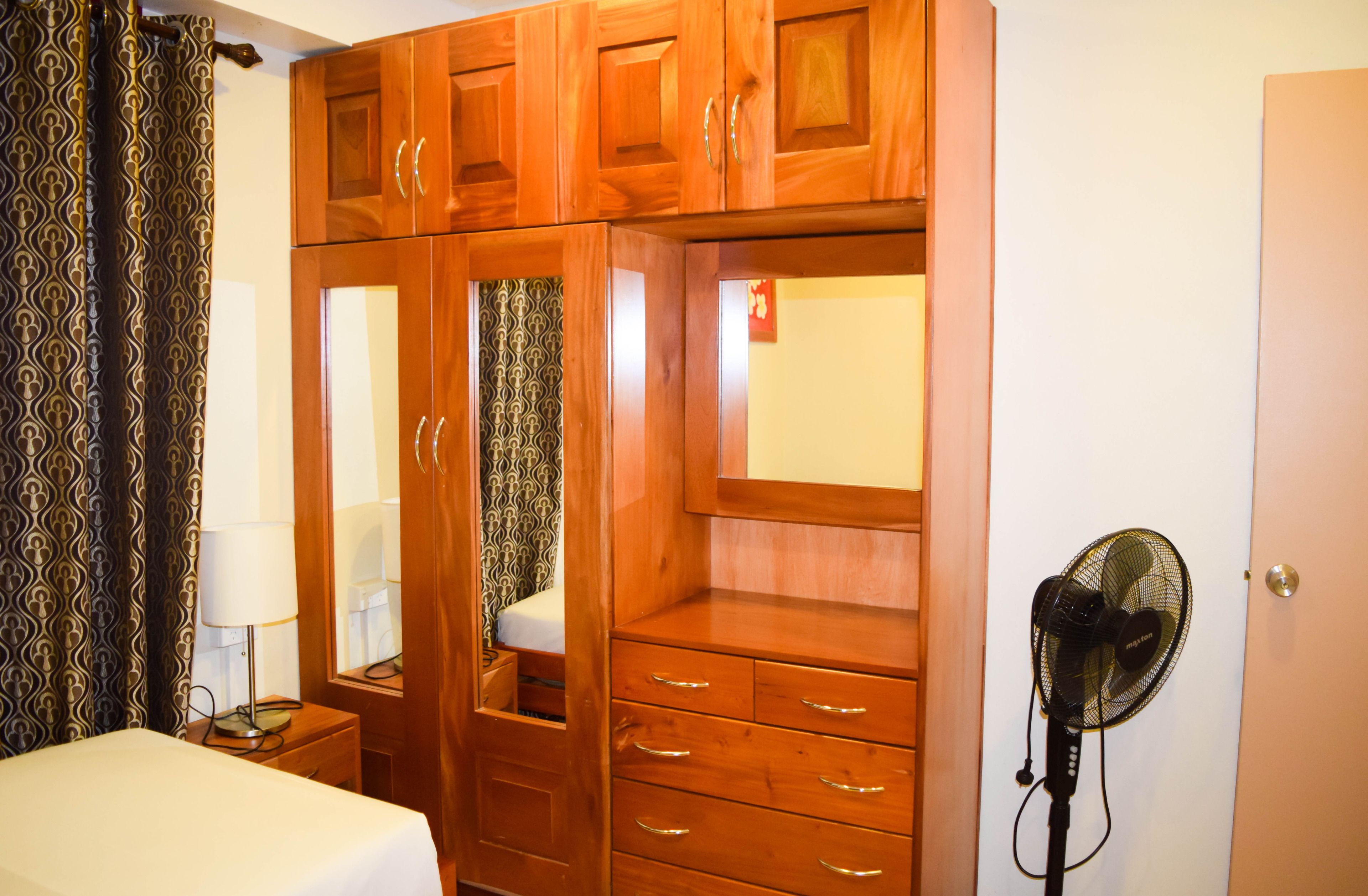 A bedroom with a wooden wardrobe, a bedside table with a lamp, and a standing fan.