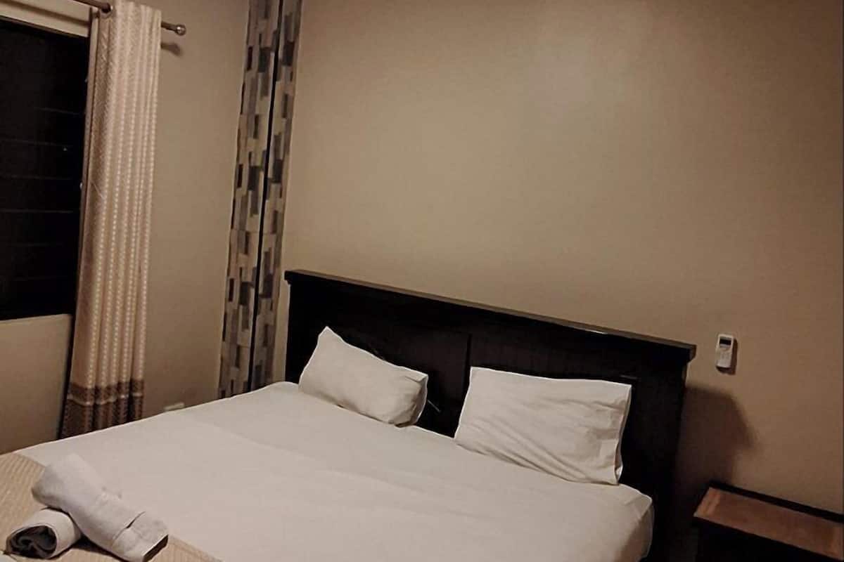 A hotel room with a large bed, a bedside table, a headboard, a wall-mounted light, and a window with curtains.