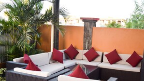 A section of outdoor seating with wicker furniture and red cushions, surrounded by greenery and a stone pillar. - Al-Minhaj Service Apartments (Nadi)