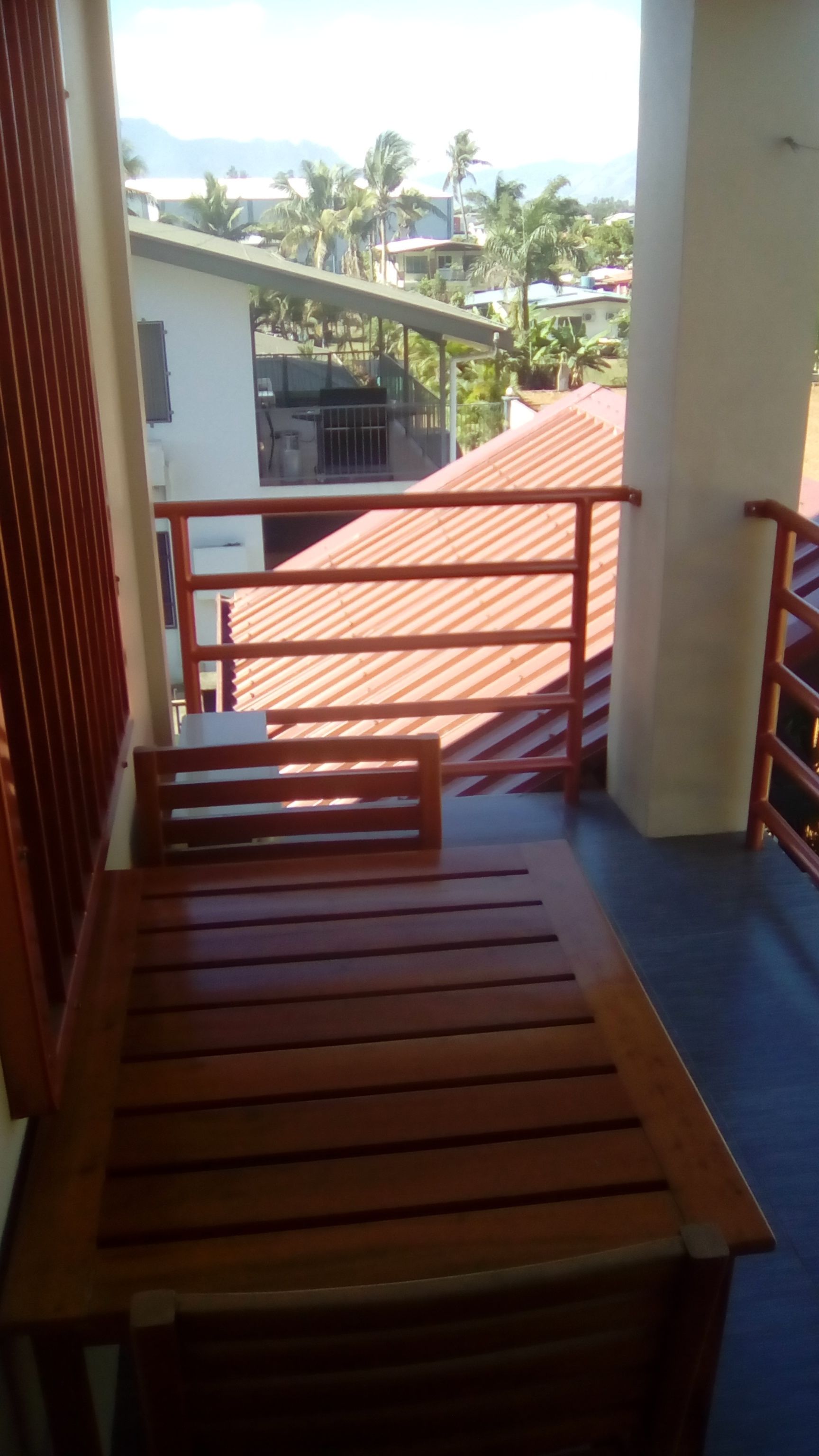 A wooden bench on a balcony with a view of a building and palm trees.