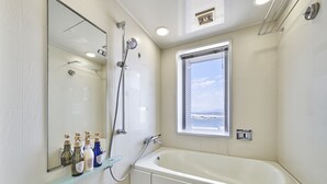 A bathroom with a bathtub, a shower, a glass shelf with toiletries, a window with a view, and a mirror.