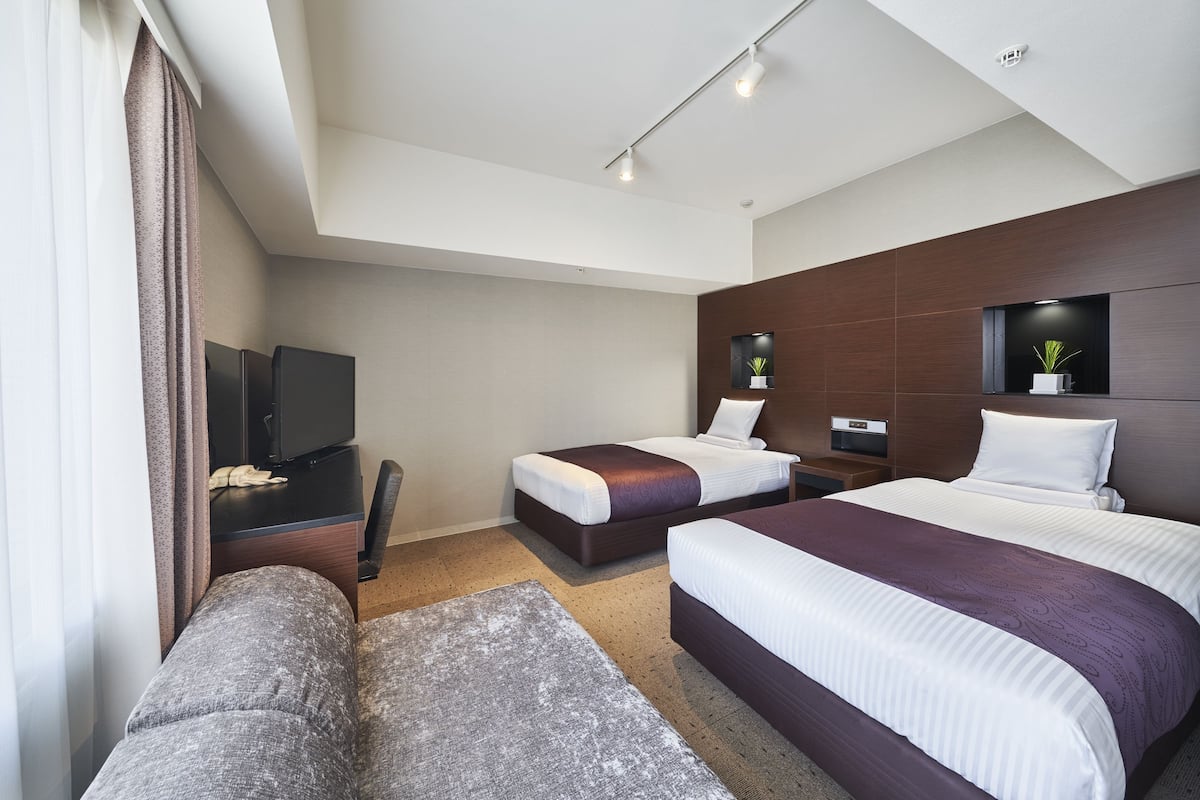 A modern hotel room with two beds, a sofa, a TV, and a small plant.