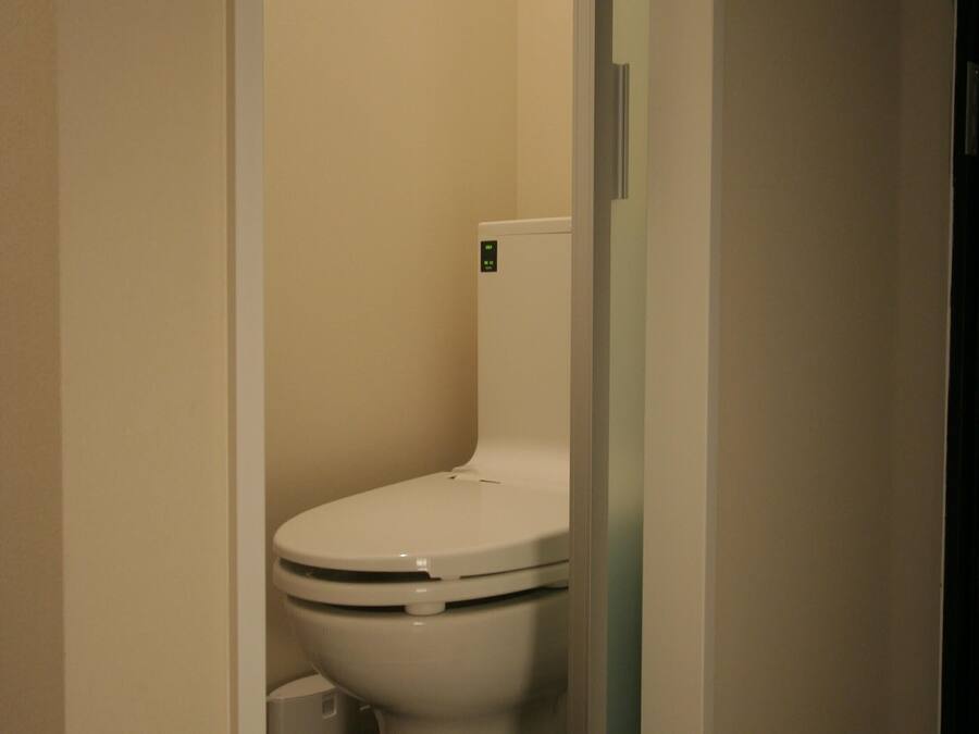 A small, narrow bathroom with a white toilet, a white trash bin, and a door leading to another room.