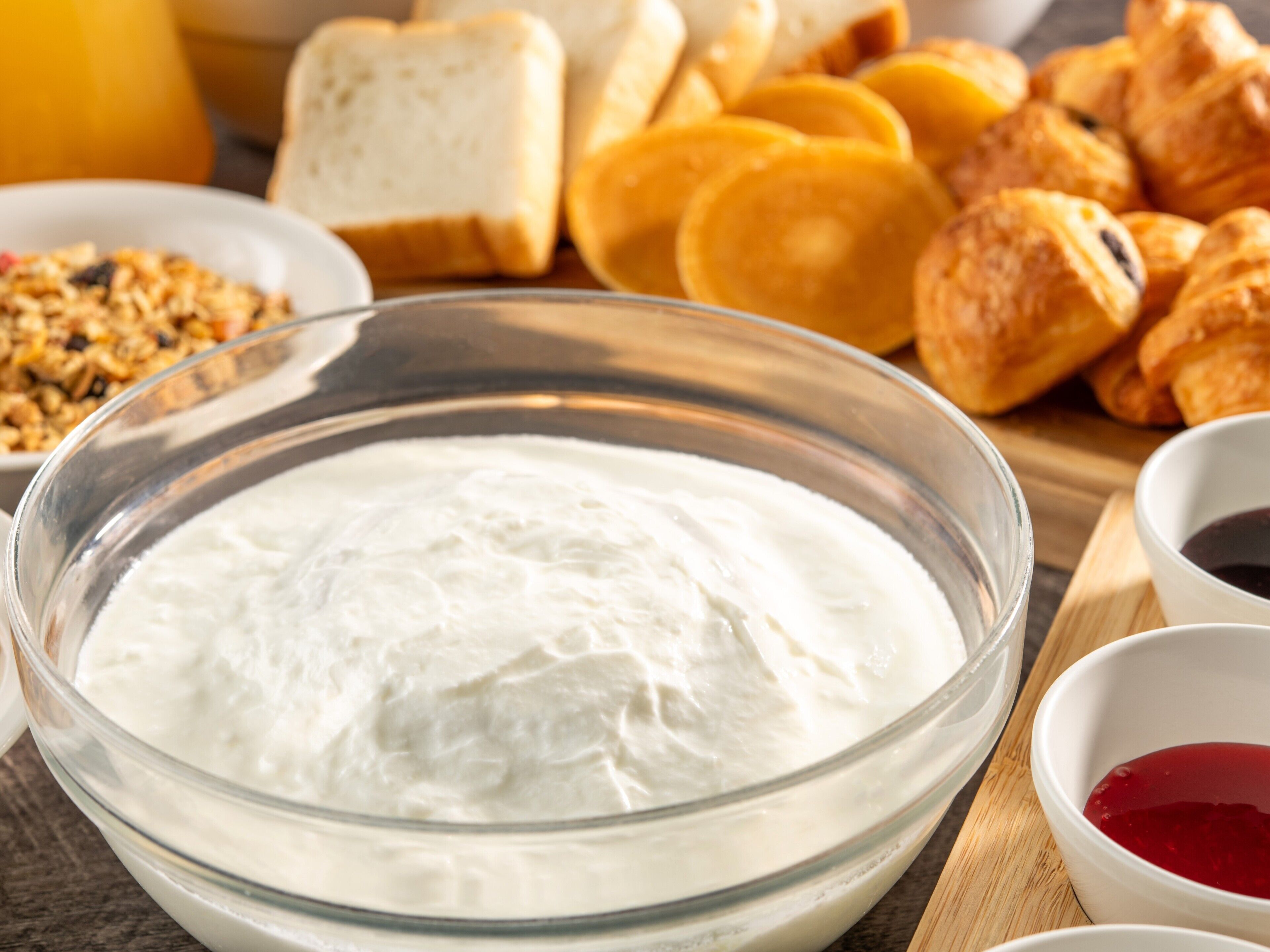A bowl of white creamy substance, possibly yogurt or sour cream, surrounded by bread and pastries on a wooden board.