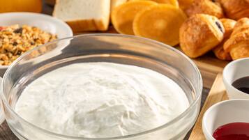 A bowl of white creamy substance, possibly yogurt or sour cream, surrounded by bread and pastries on a wooden board.