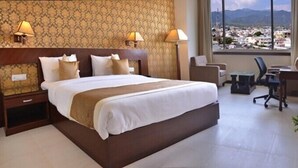 Een hotelkamer met een groot bed, twee nachtkastjes, een bureau met stoel, en uitzicht op een stadsgezicht door grote ramen.