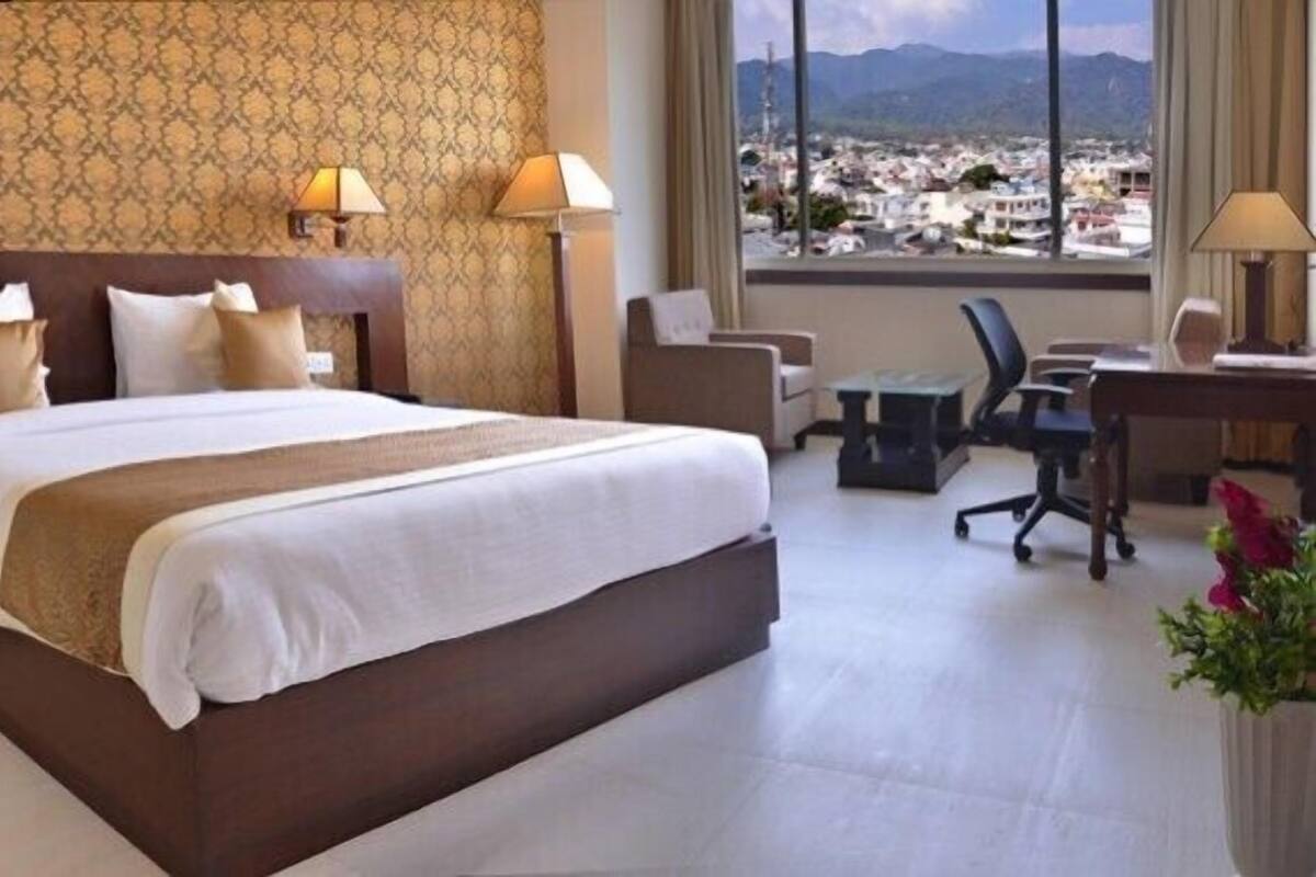 A hotel room with a large bed, two bedside tables, a desk with a chair, and a view of a cityscape through large windows.