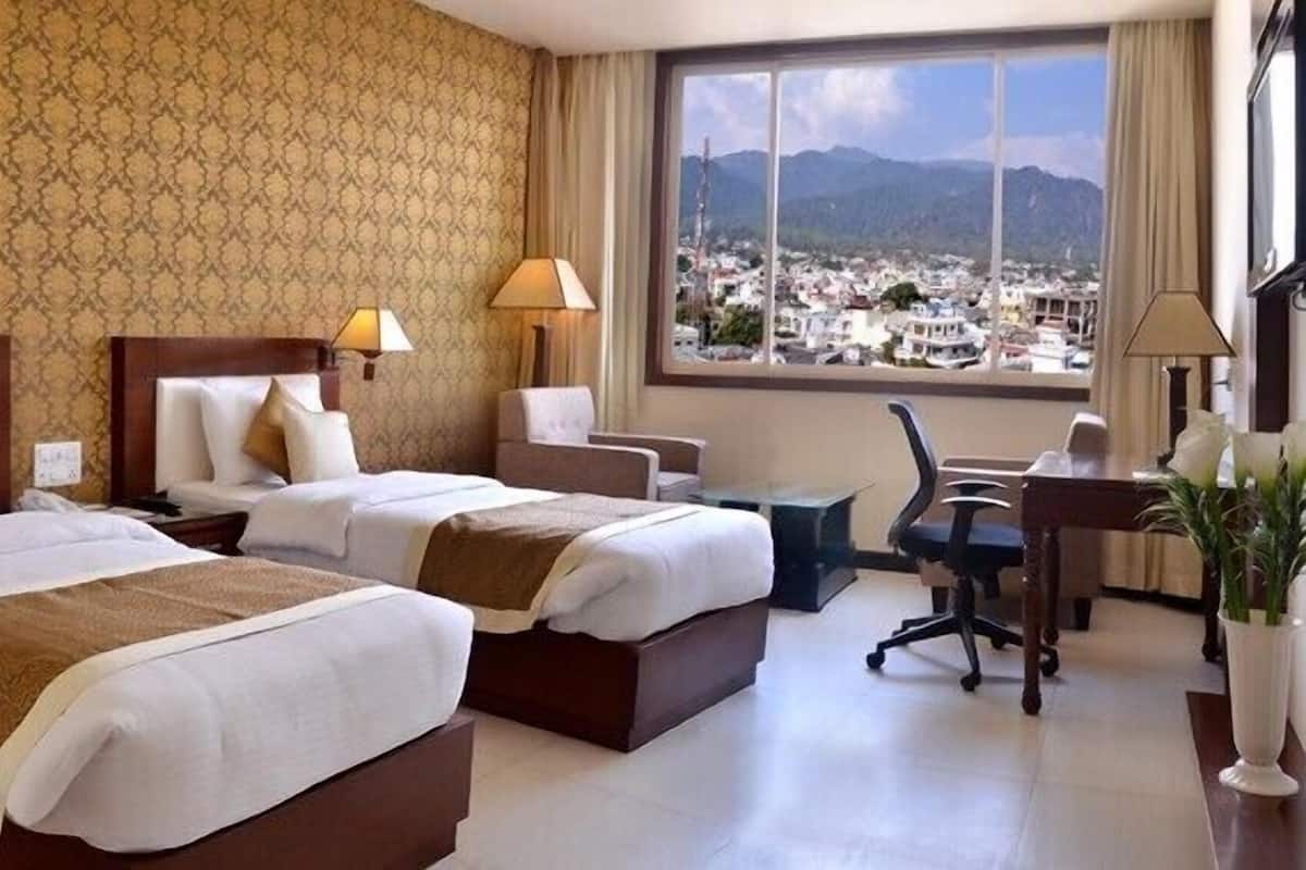 A hotel room with two beds, a desk, and a chair. There is a large window with a view of a city and mountains.
