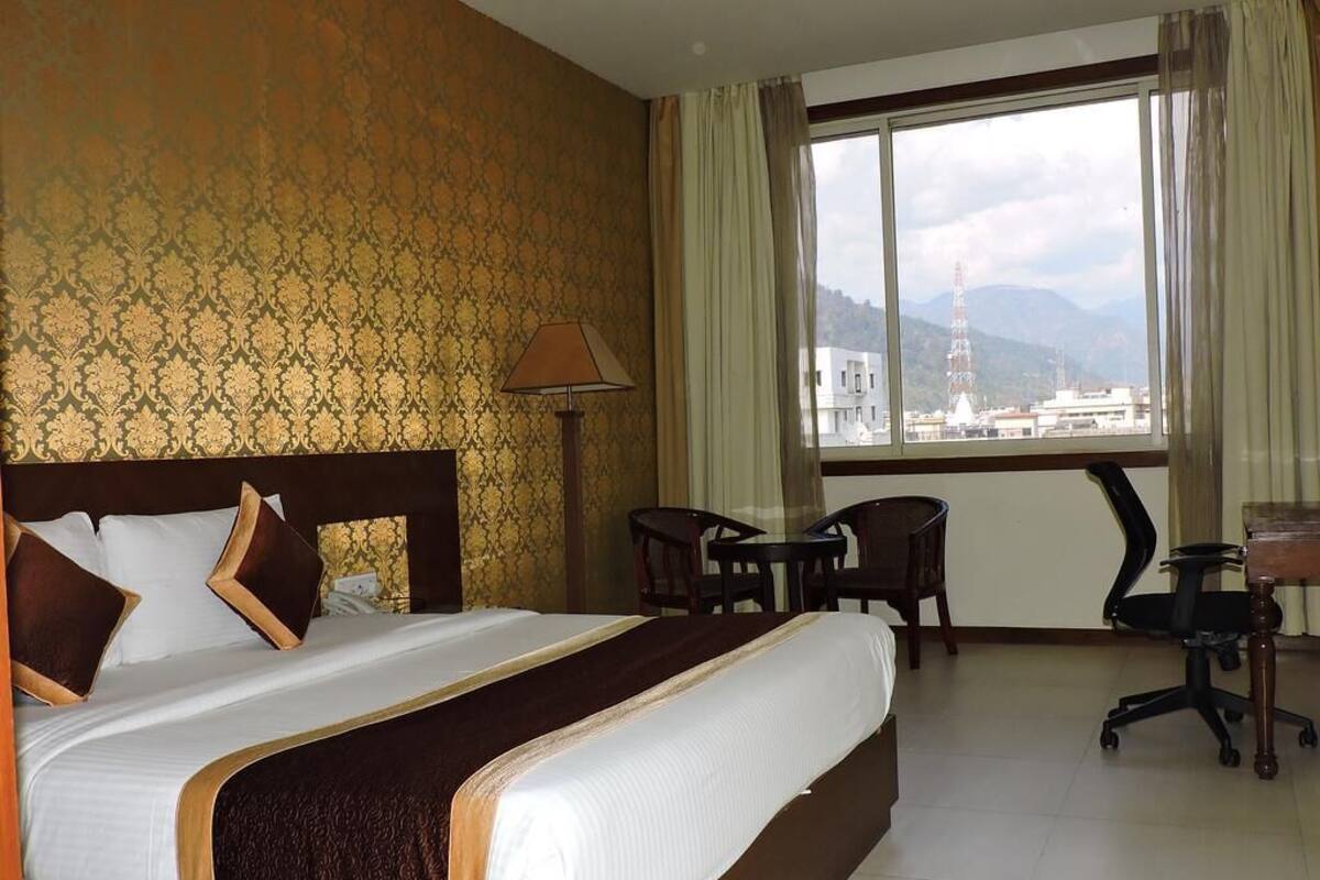 A hotel room with a large bed, a desk, two chairs, and a view of a cityscape through a large window.