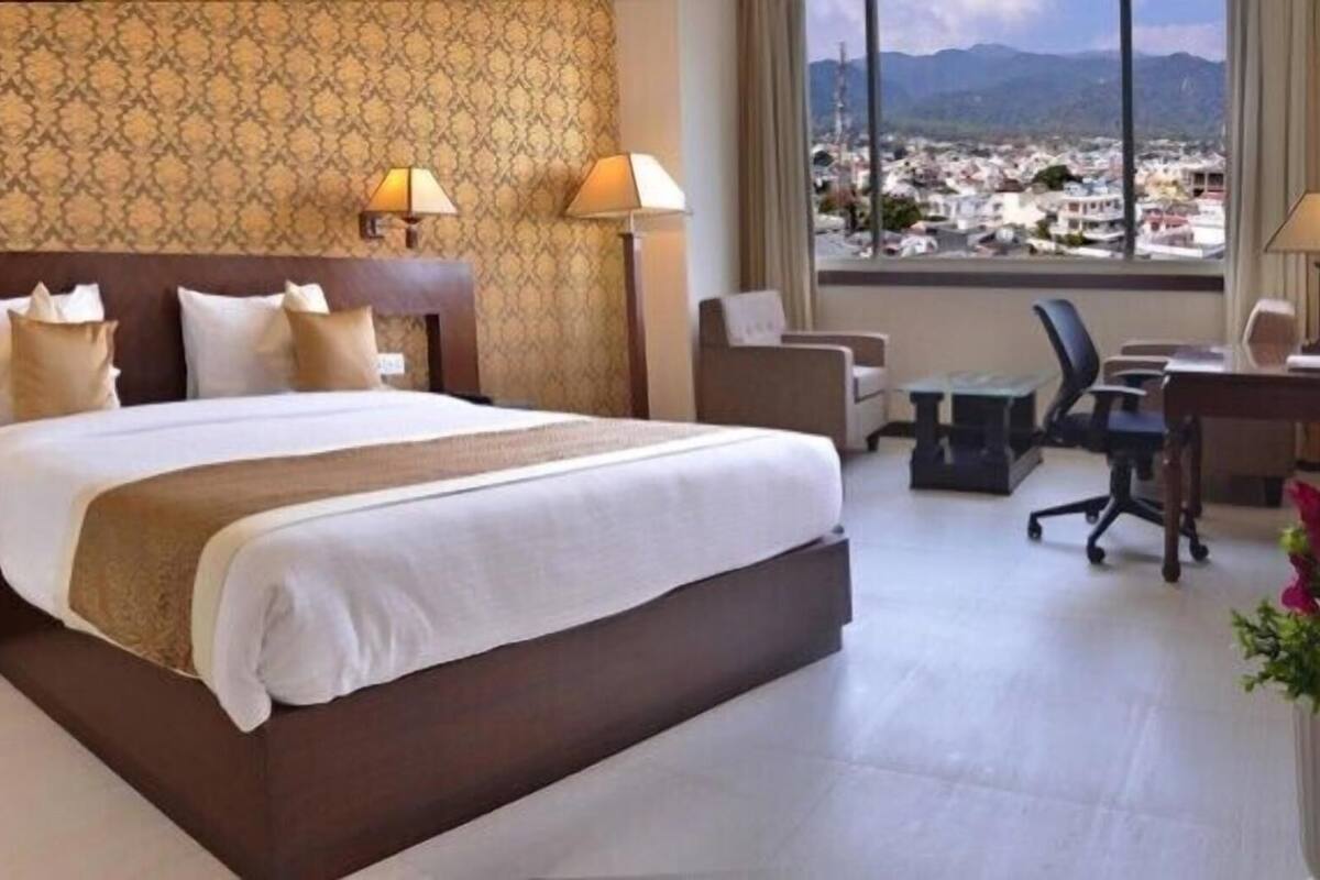 A hotel room with a large bed, two bedside tables, a desk, and a chair. There are lamps on the bedside tables and a view of a cityscape through the window.